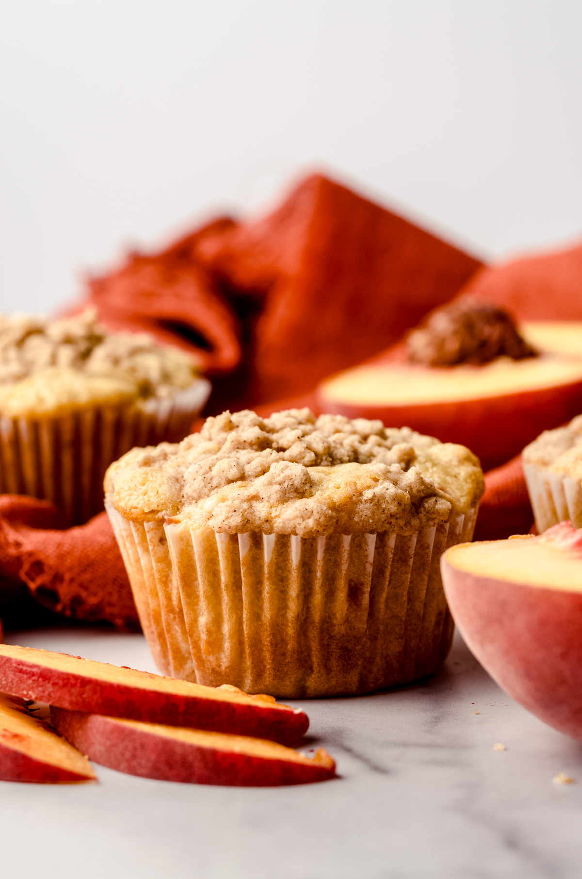 A peach muffin on a surface with an orange kitchen towel in the background with peach slices around it.