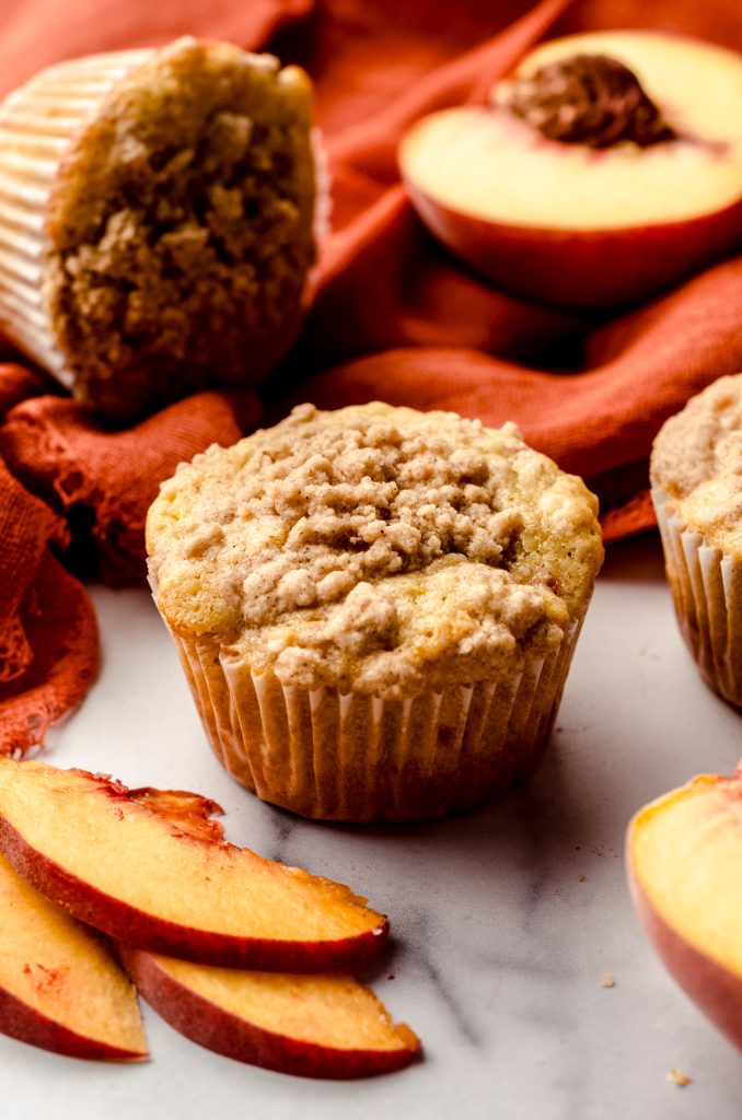 A peach muffin on a surface with an orange kitchen towel in the background with peach slices around it.