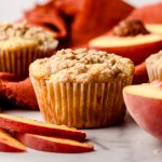 A peach muffin on a surface with an orange kitchen towel in the background with peach slices around it.