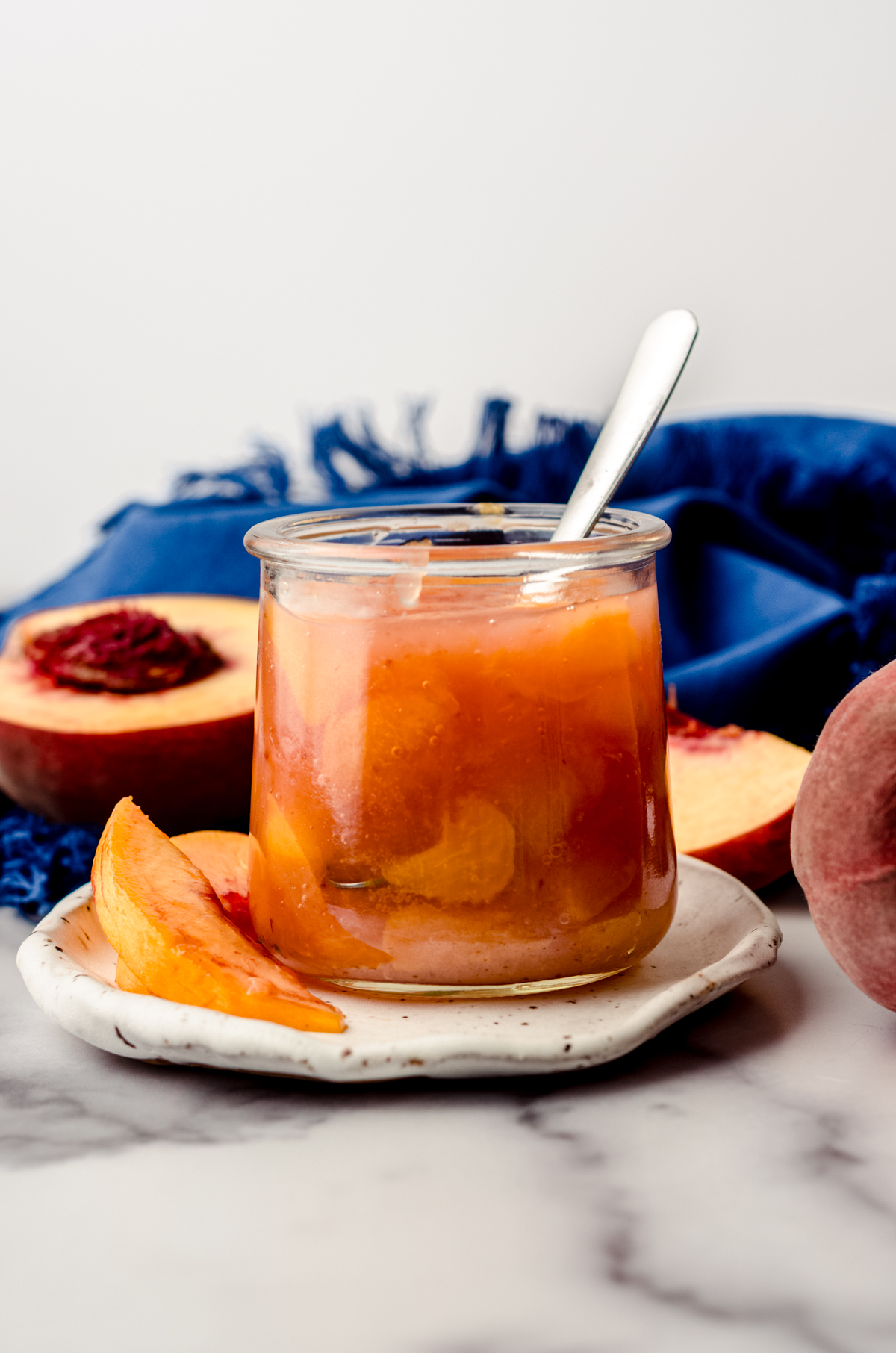 A jar of peach compote sitting on a plate with peach slices and a small spoon.