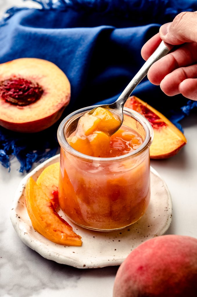 Someone is using a small spoon to scoop out of a jar of peach compote sitting on a plate with peach slices.