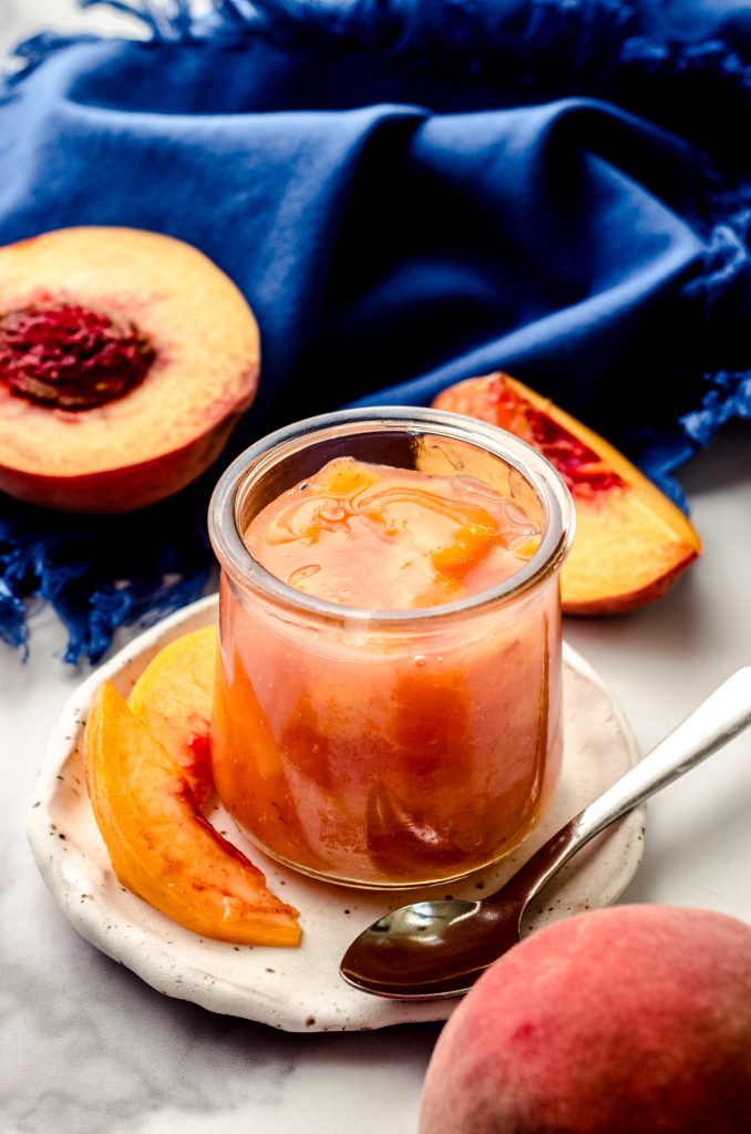 A jar of peach compote sitting on a plate with peach slices and a small spoon.