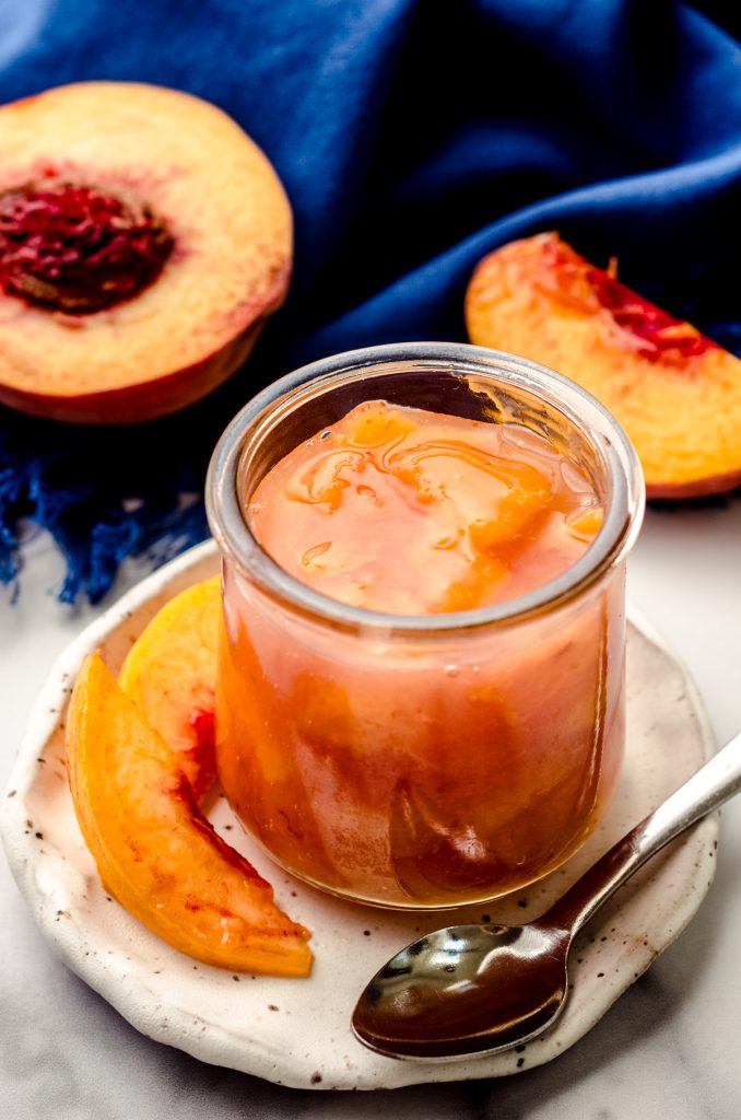 A jar of peach compote sitting on a plate with peach slices and a small spoon.