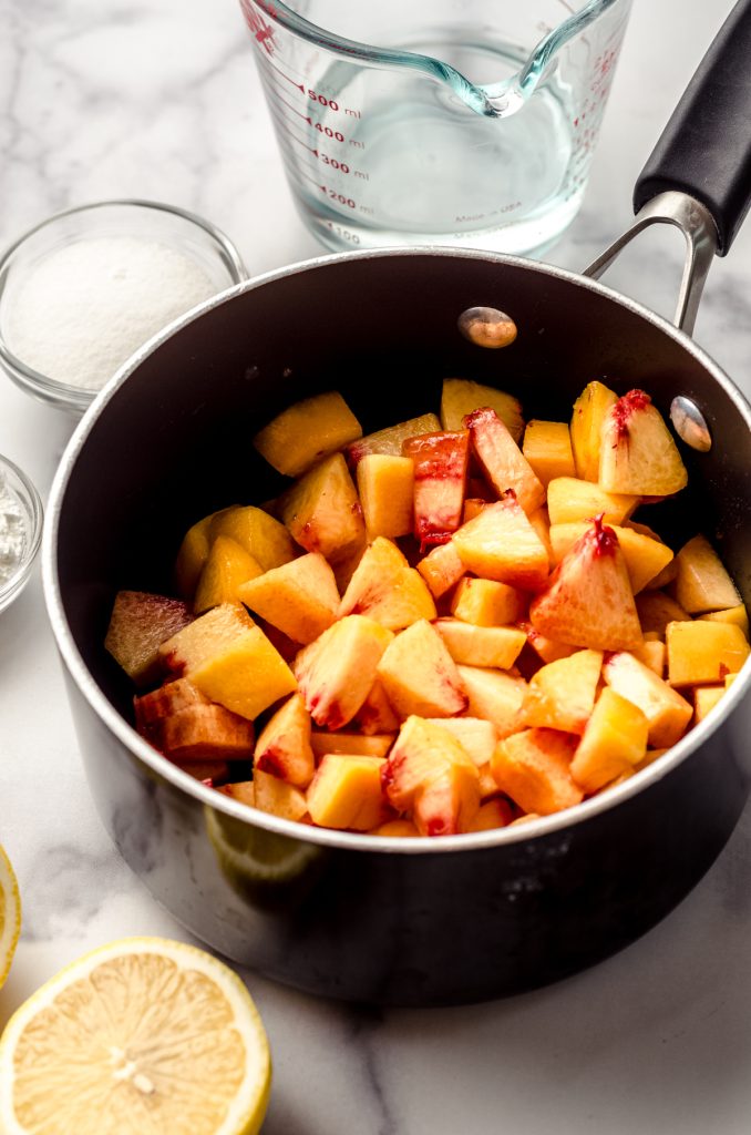 Chopped peaches in a saucepan with a lemon half and a bowl of sugar around it.