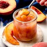 Someone is using a small spoon to scoop out of a jar of peach compote sitting on a plate with peach slices.