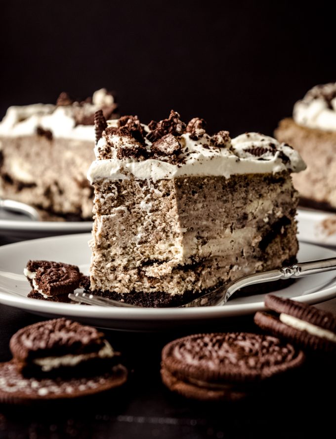 A slice of cookies and cream cheesecake on a plate with a fork and Oreo chunks and a bite has been taken out of it.