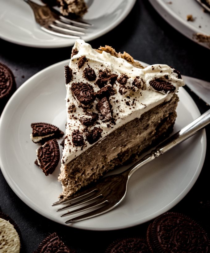 A slice of cookies and cream cheesecake on a plate with a fork and Oreo chunks.