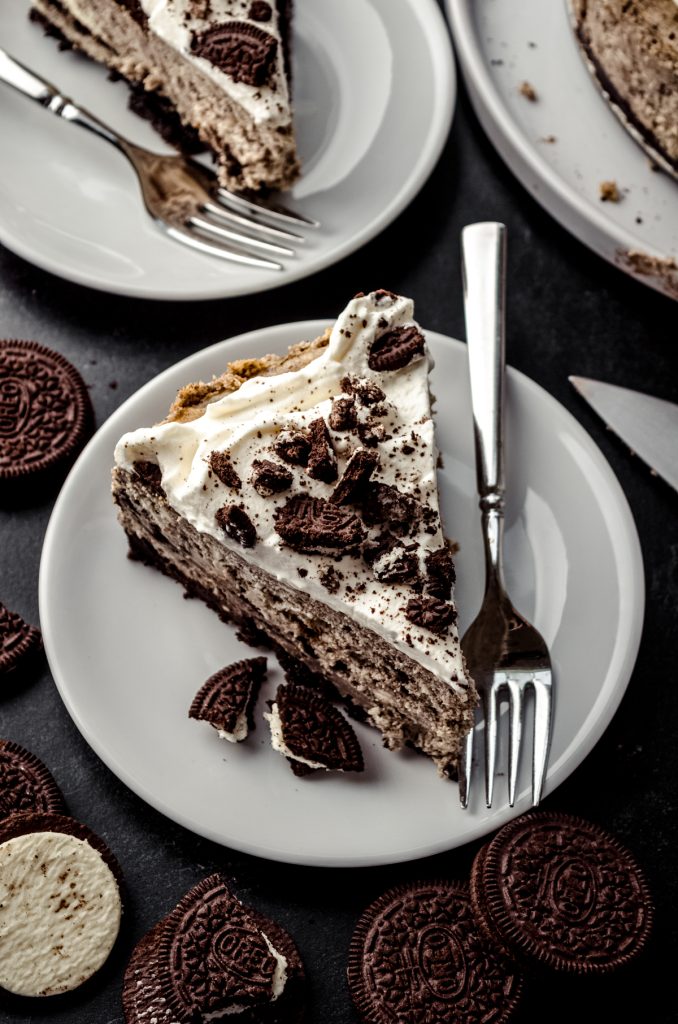 A slice of cookies and cream cheesecake on a plate with a fork and Oreo chunks.