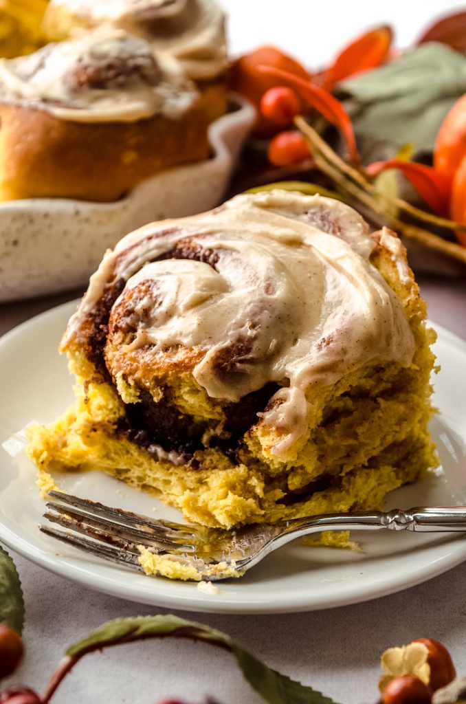 A pumpkin cinnamon roll on a plate with a fork and a bite taken out of it.