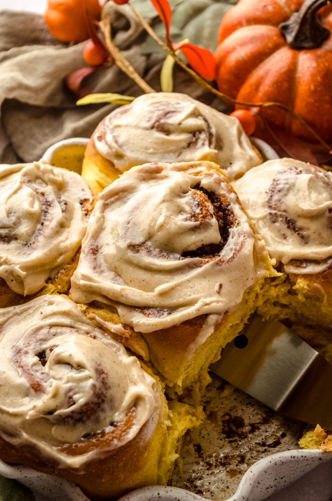 Someone is using a spatula to lift a pumpkin cinnamon roll topped with maple cinnamon cream cheese frosting out of a baking dish.