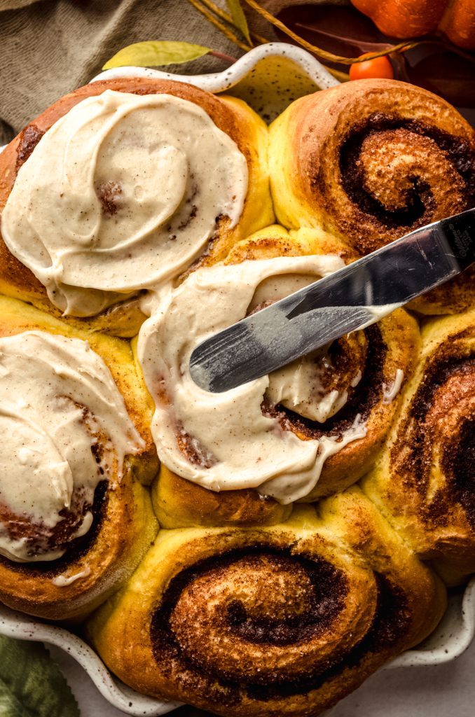 Someone is using a spatula to spread maple cinnamon cream cheese frosting onto baked pumpkin cinnamon rolls.