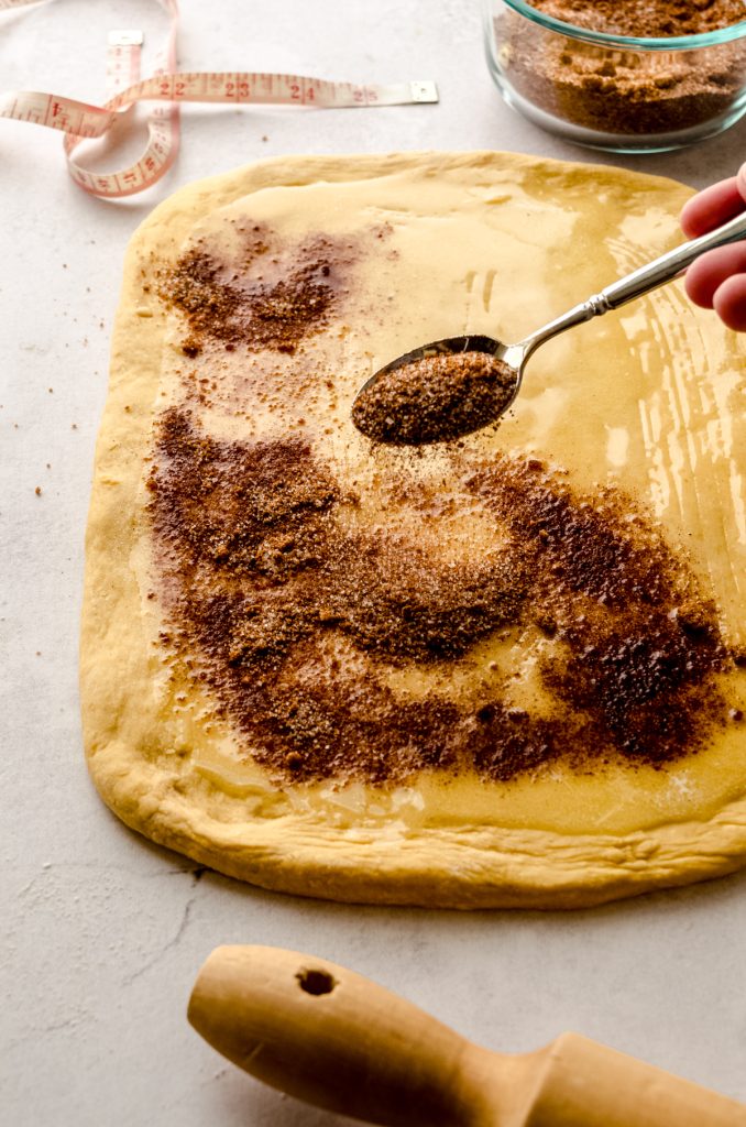 Someone is sprinkling cinnamon sugar onto a rectangle of dough to make pumpkin cinnamon rolls.