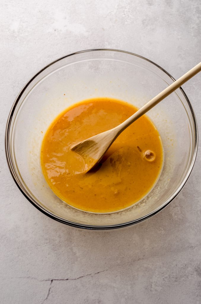 A mixture of pumpkin and other wet ingredients to make pumpkin cinnamon rolls in a large bowl with a wooden spoon.