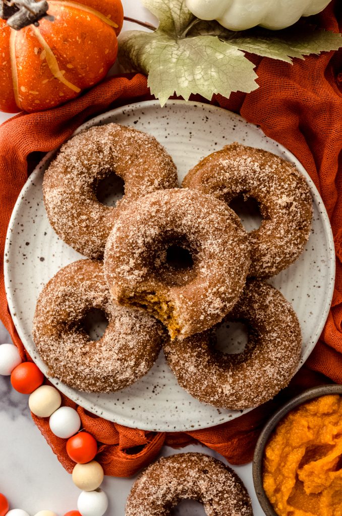 Aerial photo of a stack of baked pumpkin donuts on a plate and someone is holding the one on the top that has a bite taken out of it.