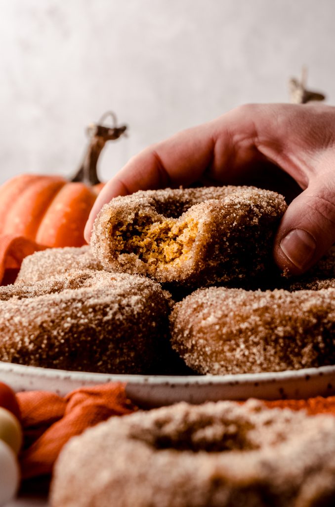 A photo of a stack of baked pumpkin donuts on a plate and someone is holding the one on the top that has a bite taken out of it.