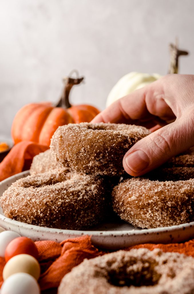 A plate of baked pumpkin donuts and someone is holding the one on the top of the stack.