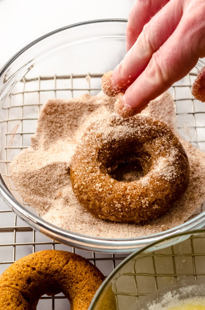 Someone is dunking a baked pumpkin donut into a bowl of spiced sugar coating.