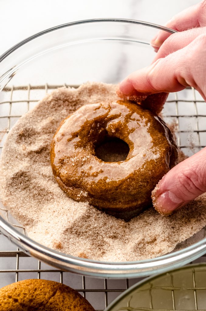 Someone is dunking a baked pumpkin donut into a bowl of spiced sugar coating.