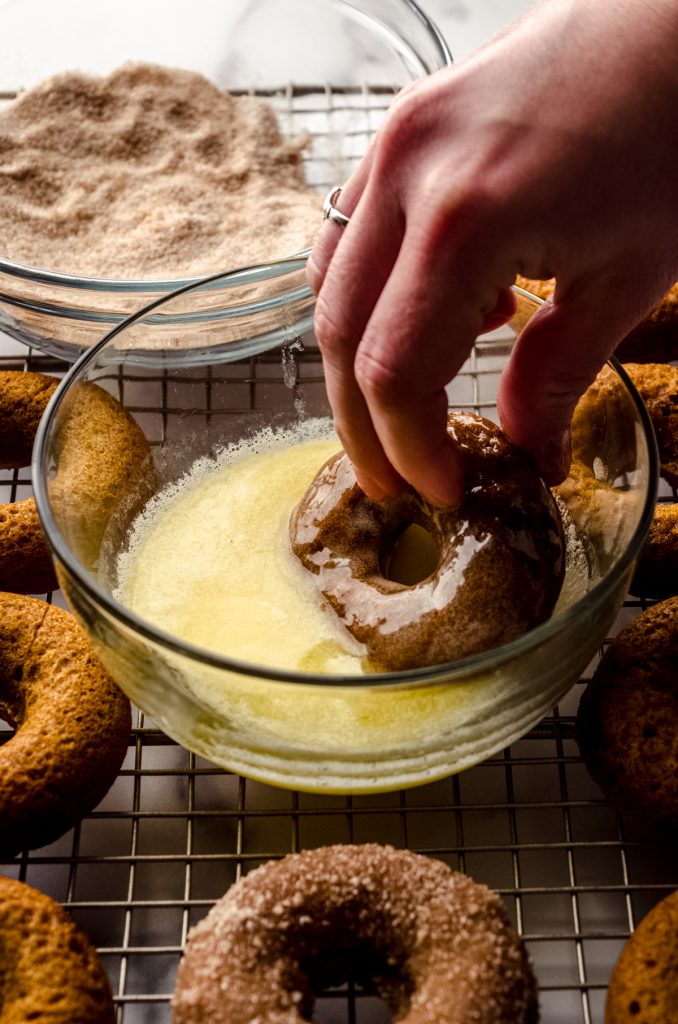 Someone is dunking a baked pumpkin donut into a bowl of melted butter before putting on a spiced sugar coating.