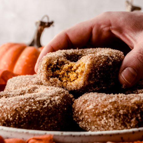 A photo of a stack of baked pumpkin donuts on a plate and someone is holding the one on the top that has a bite taken out of it.