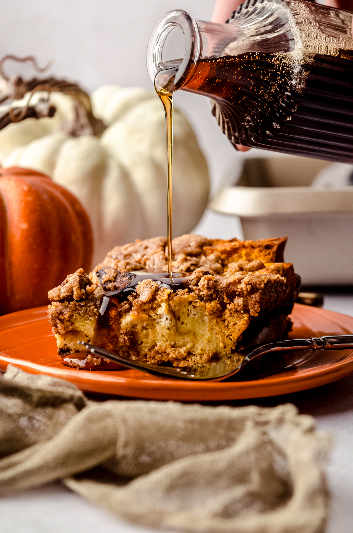 A slice of pumpkin French toast casserole on a plate and someone is pouring syrup onto it.