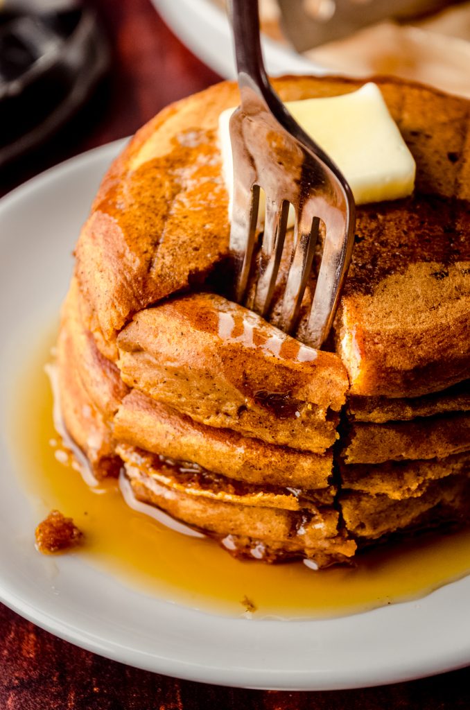 A stack of pumpkin pancakes with pats of butter and syrup on it that has been sliced into a triangle shaped stack of bites and a fork is stuck in the bite.