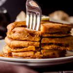 A stack of pumpkin pancakes with pats of butter and syrup on it that has been sliced into a triangle shaped stack of bites and a fork is stuck in the bite.