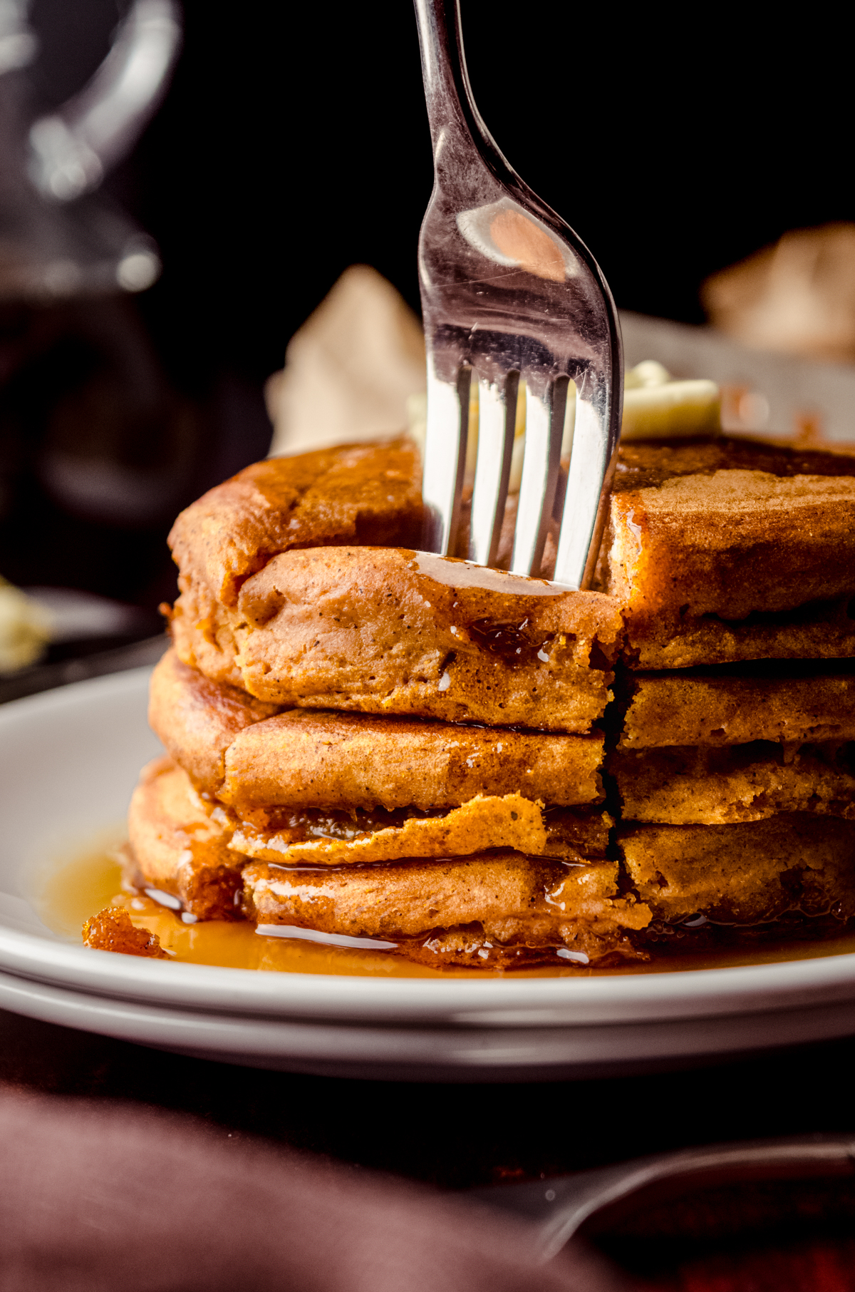 A stack of pumpkin pancakes with pats of butter and syrup on it that has been sliced into a triangle shaped stack of bites and a fork is stuck in the bite.