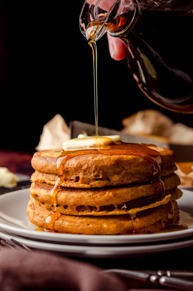 A small stack of pumpkin pancakes with pats of butter on top and someone is pouring syrup onto it.