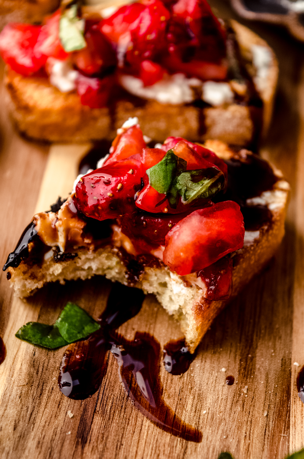 A slice of strawberry goat cheese bruschetta on a wooden cutting board with a bite taken out of it.