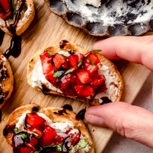 Aerial photo of strawberry goat cheese bruschetta pieces on a wooden cutting board and someone is grabbing one of the slices with their hand.