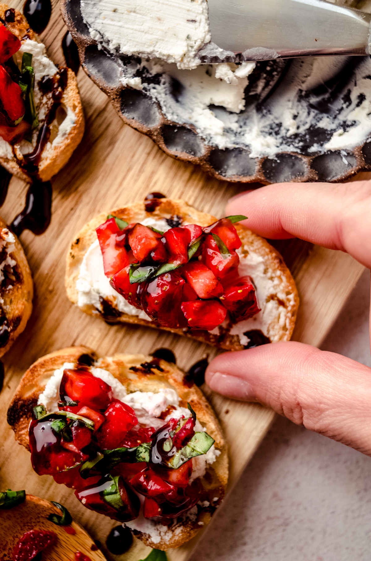 Aerial photo of strawberry goat cheese bruschetta pieces on a wooden cutting board and someone is grabbing one of the slices with their hand.