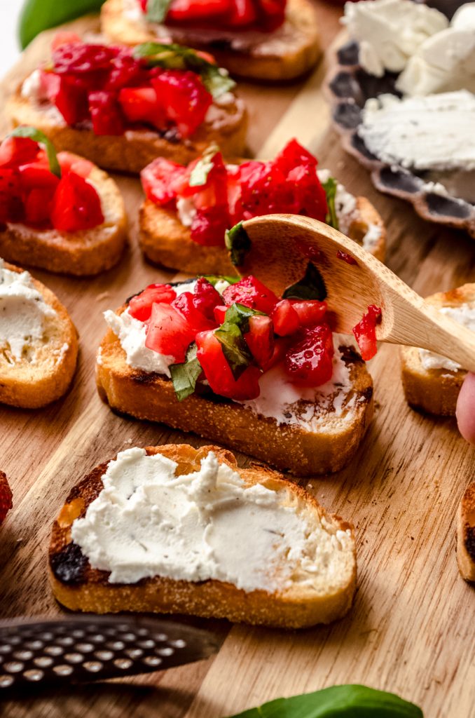 Someone is spooning a strawberry and basil mixture onto grilled French bread slices that have been spread with goat cheese to make strawberry bruschetta.