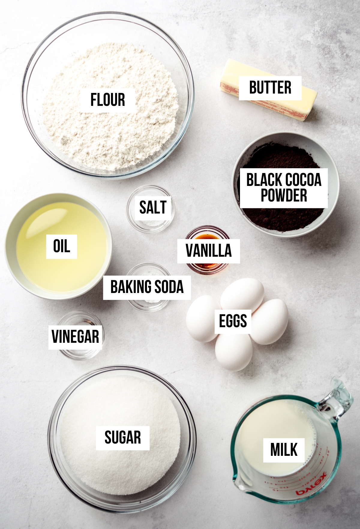 Aerial photo of ingredients to make black cocoa chocolate cake with text overlay labeling each ingredient.