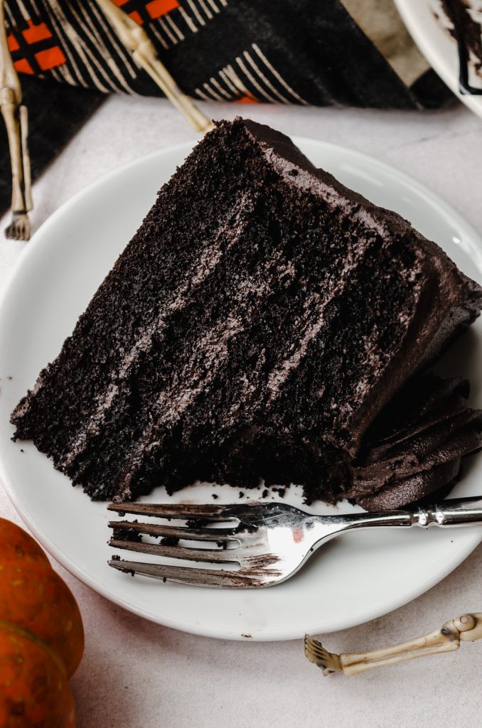 A slice of black cocoa chocolate cake on a plate with a fork and a bite has been taken out of the cake.