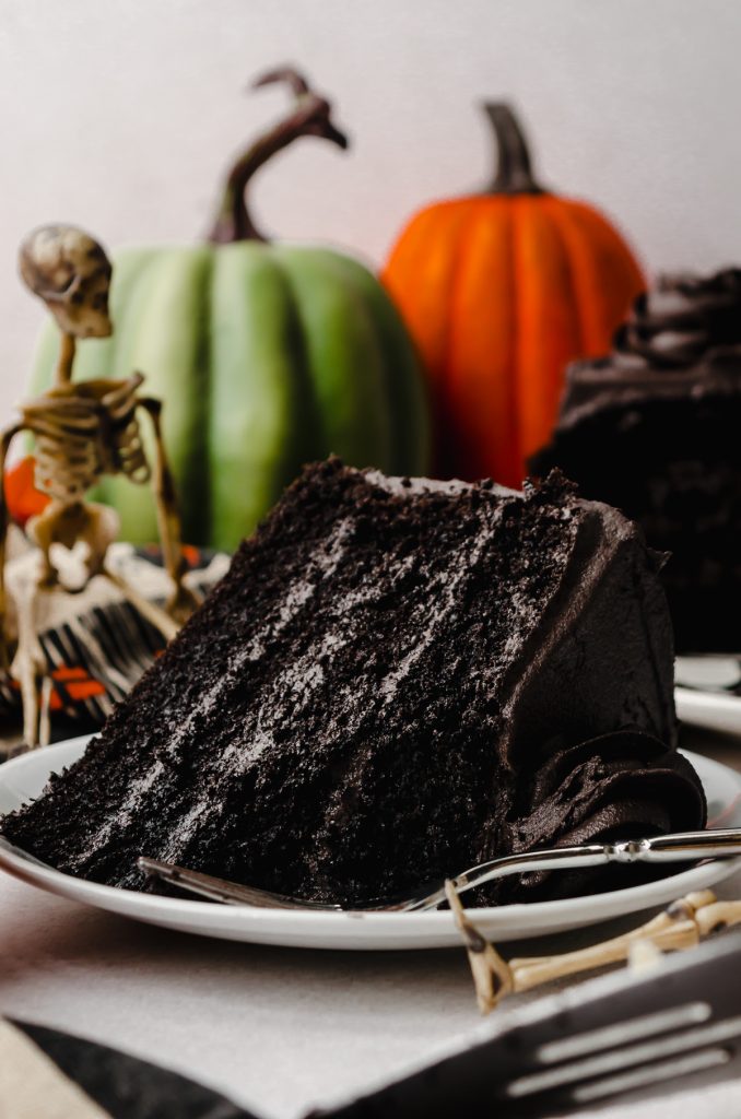 A slice of black cocoa chocolate cake on a plate with green and orange pumpkins and a skeleton on the background and a fork on the plate and a bite has been taken out of the cake.