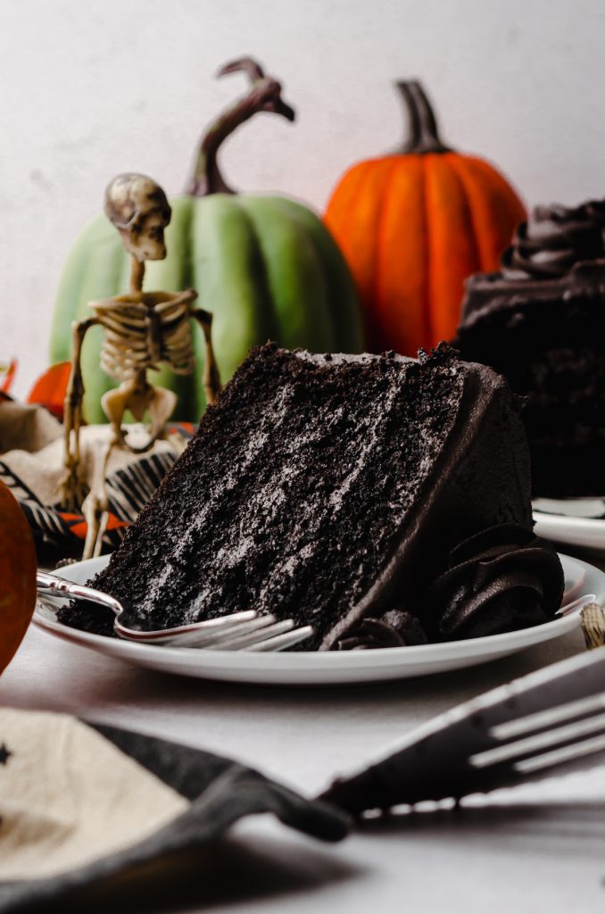 A slice of black cocoa chocolate cake on a plate with green and orange pumpkins and a skeleton on the background and a fork on the plate.