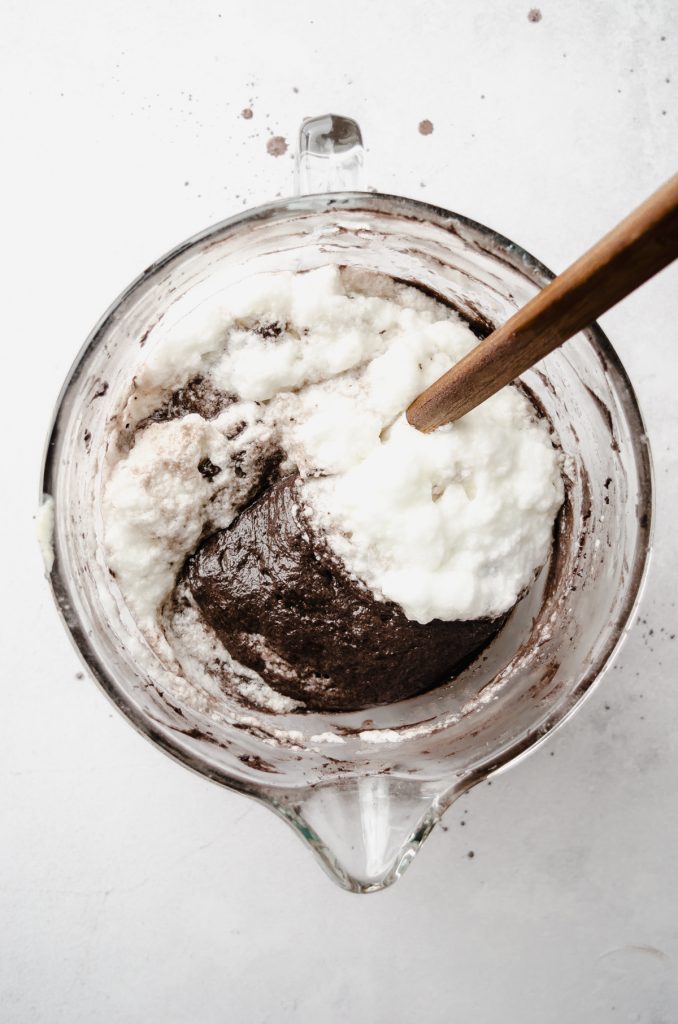 Aerial photo of batter for black cocoa chocolate cake in a glass bowl with a spatula and egg whites are being folded into the batter.