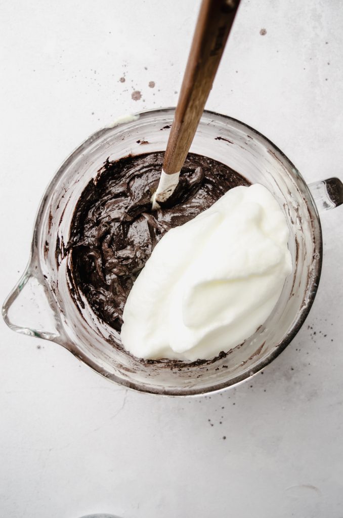 Aerial photo of batter for black cocoa chocolate cake and whipped egg whites in a glass bowl with a spatula.