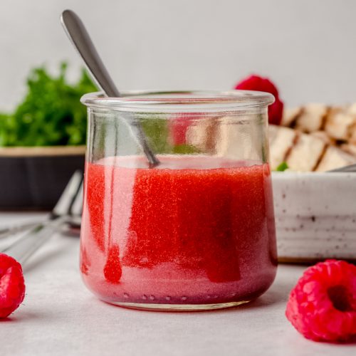 A jar of raspberry vinaigrette on a surface with a spoon in it and you can see raspberries in the foreground and salad in plates in the background.