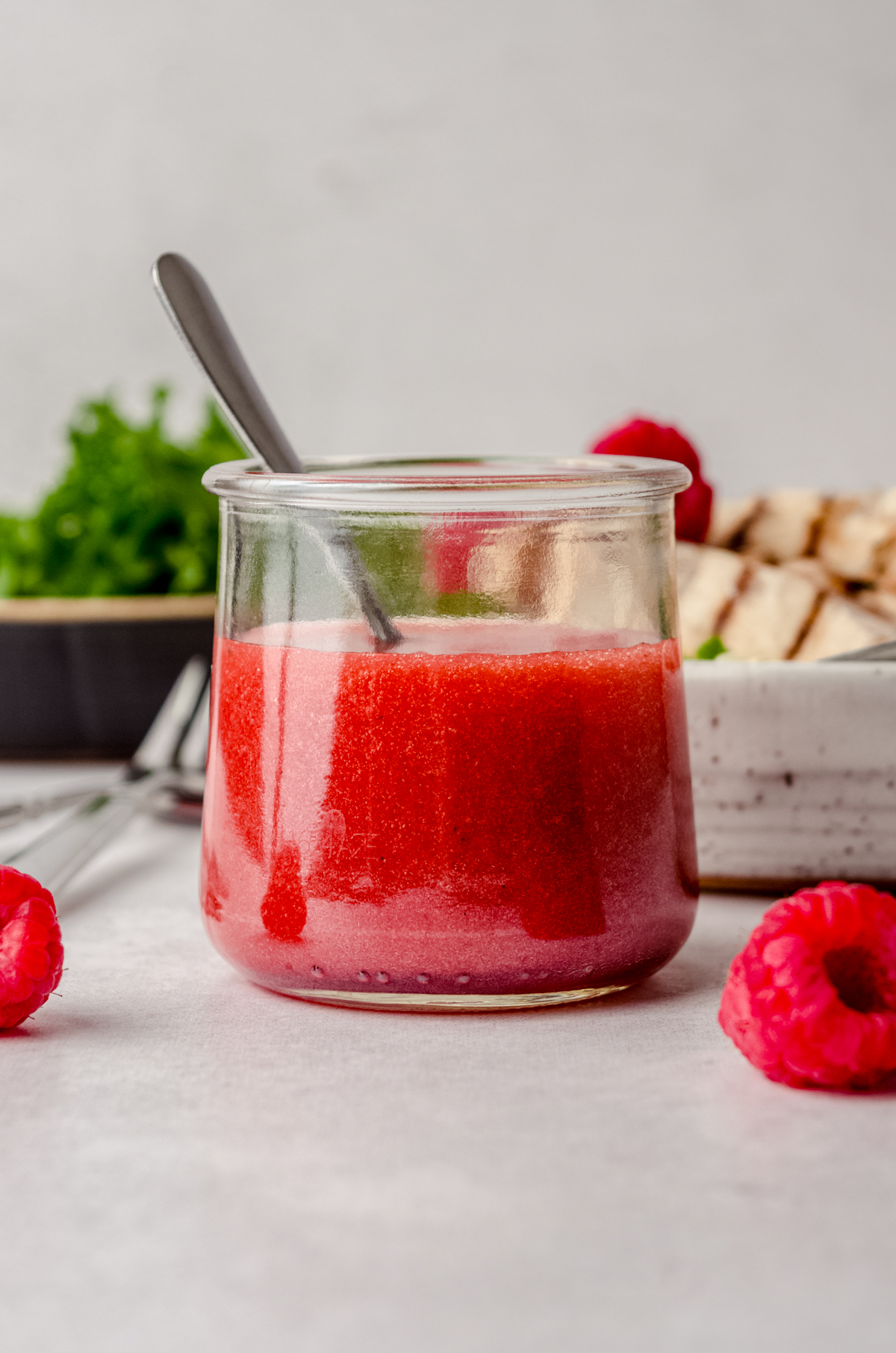 A jar of raspberry vinaigrette on a surface with a spoon in it and you can see raspberries in the foreground and salad in plates in the background.