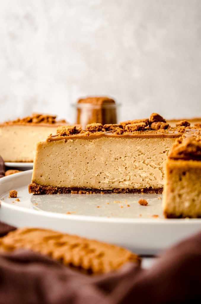 A Biscoff cheesecake that has been sliced so you can see the cross section sitting on a plate with a spatula and cup of Biscoff cookies in the background.