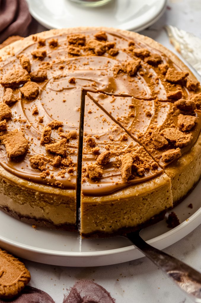 A Biscoff cheesecake on a plate and someone is using a serving utensil to remove a slice.