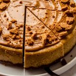 A Biscoff cheesecake on a plate and someone is using a serving utensil to remove a slice.