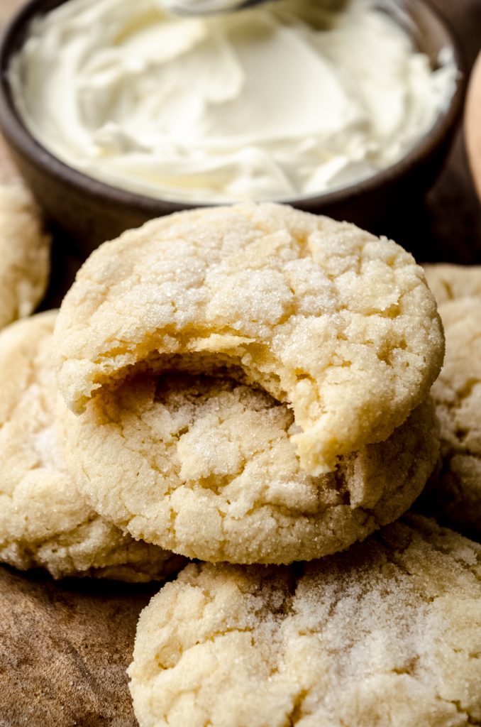 Cream cheese sugar cookies on a surface and a bite taken out of the one on the top with a bowl of cream cheese in the background.