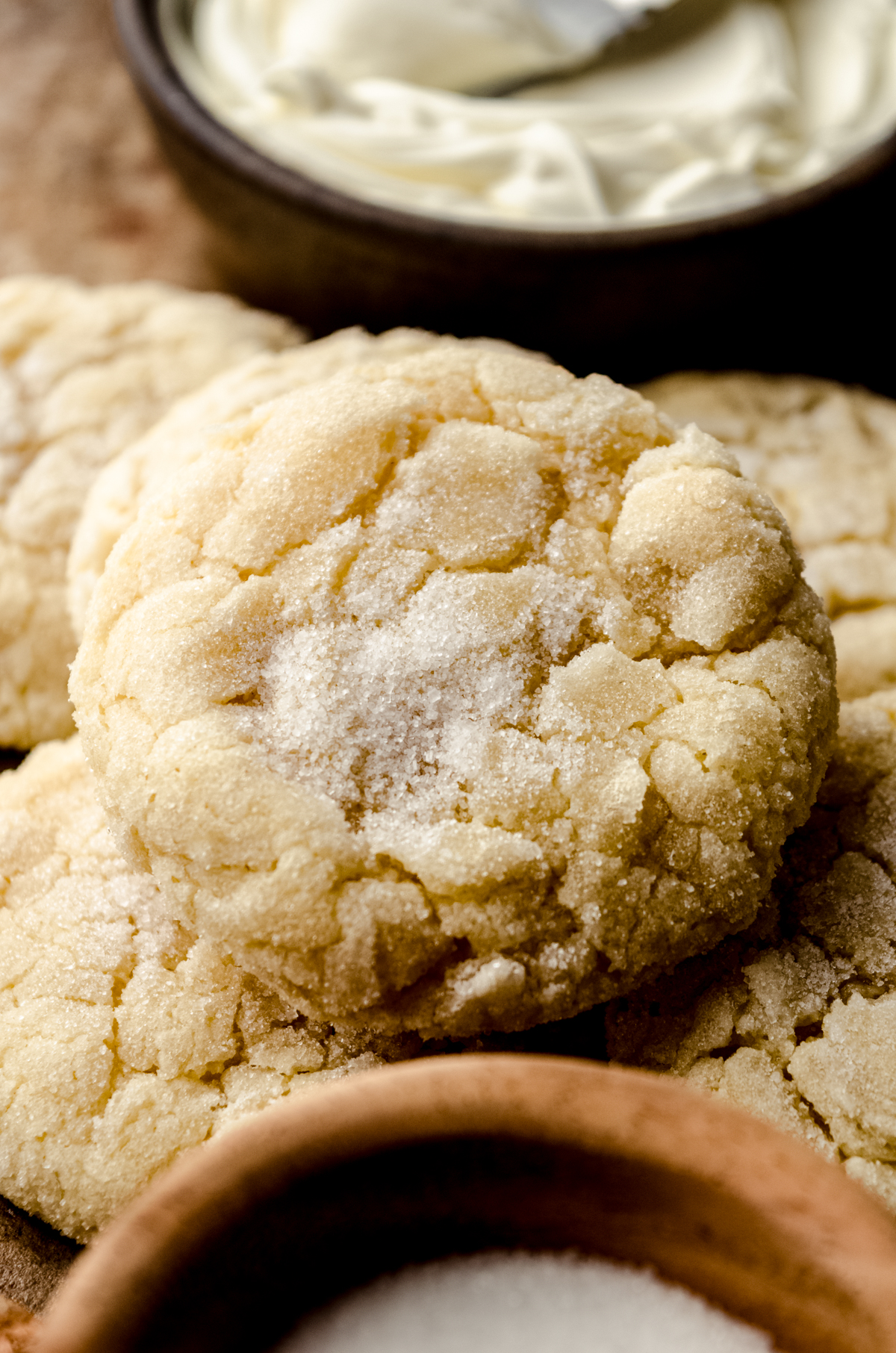 Cream cheese sugar cookies on a surface and a small bowl of sugar and a bowl of cream cheese can be seen around it.