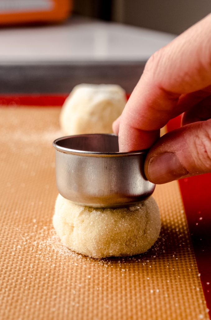 Someone is using a measuring cup to flatten a cookie dough ball on a baking sheet.