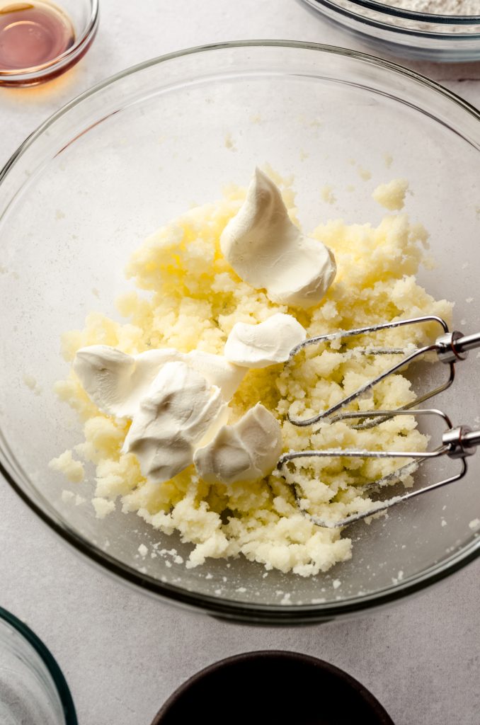 Sugar cookie dough in a bowl with chunks of cream cheese and electric beaters getting ready to mix together to make cream cheese sugar cookie dough.