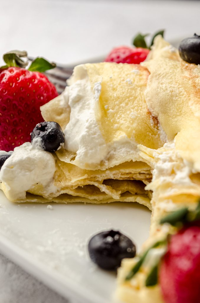 A stack of crepes with fresh whipped cream, blueberries, and strawberries and a bite has been taken out so you can see the cross section of the crepes.