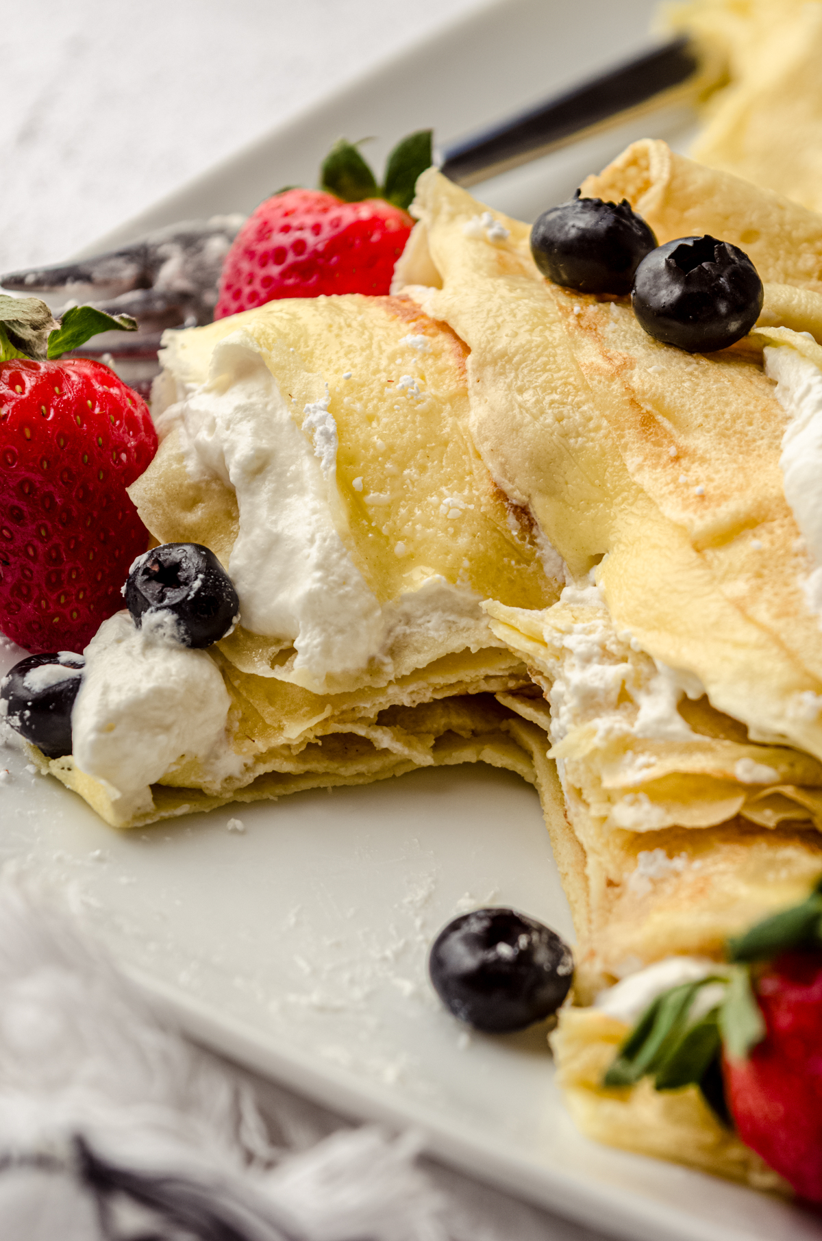 A stack of crepes with fresh whipped cream, blueberries, and strawberries and a bite has been taken out so you can see the cross section of the crepes.